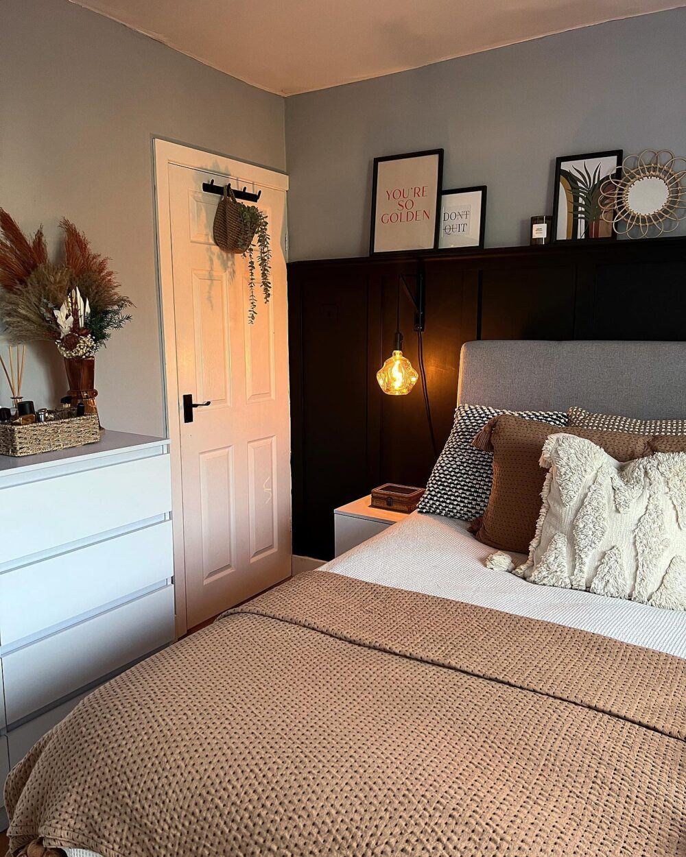 A bedroom filled with earthy and grounded tones, natural flaire with plants spread about, and a half-panel shelf above a bed that displays pictures and candles alike.